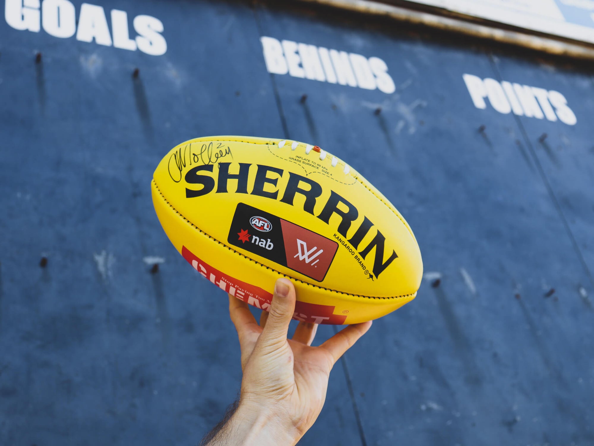 Holding a Sherrin AFL football in front of a score board.